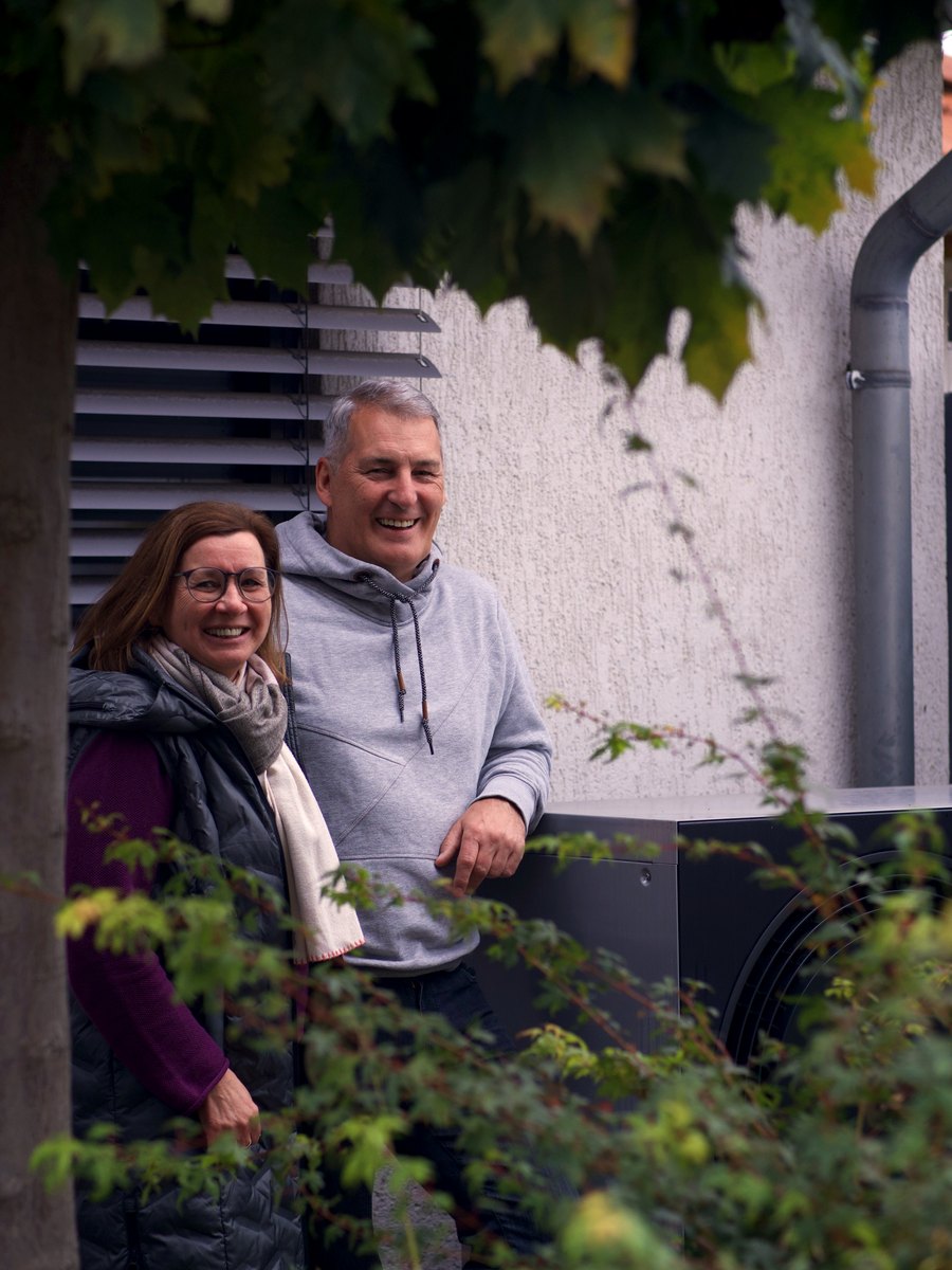 Kathi und Jörg stehen lächelnd neben ihrer neuen Wärmepumpe im Garten