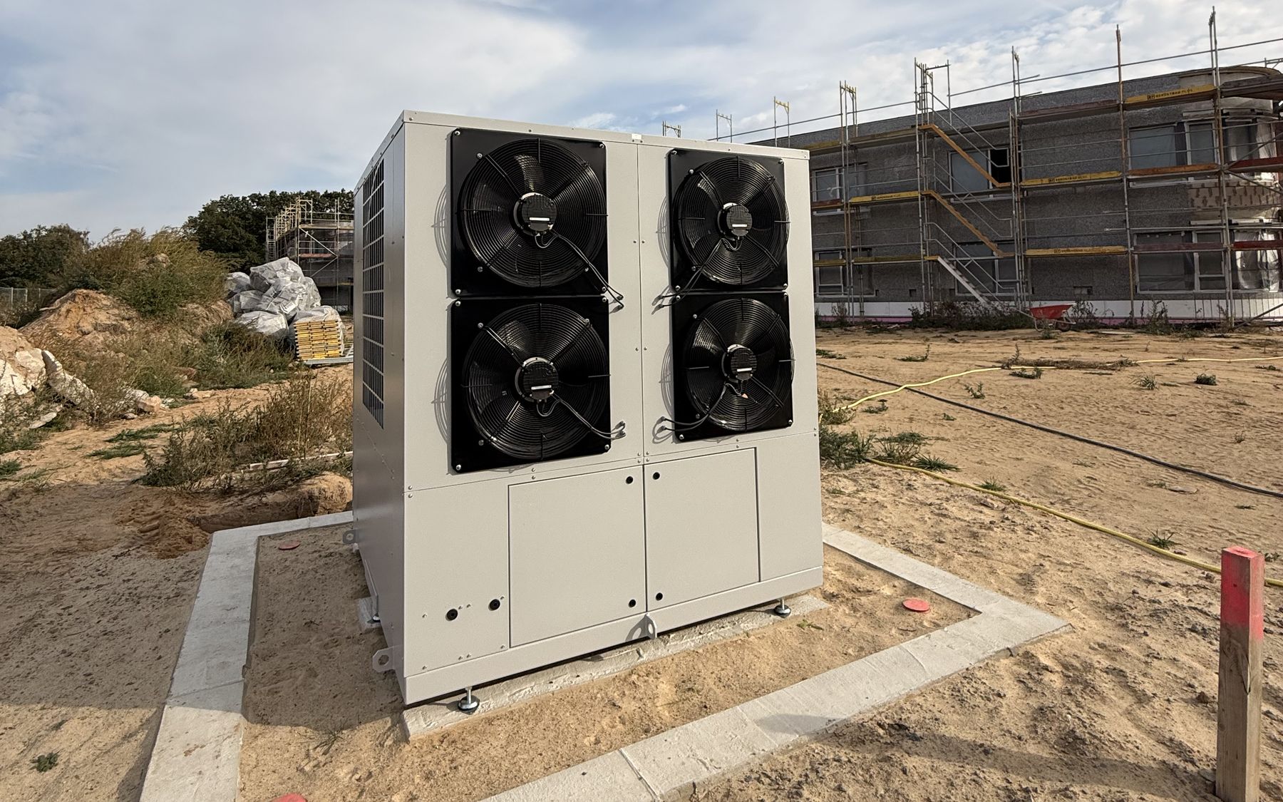 ATEC-Großwärmepumpe auf ihrem Fundament, dahinter der Rohbau des Wohnheims mit Gerüst